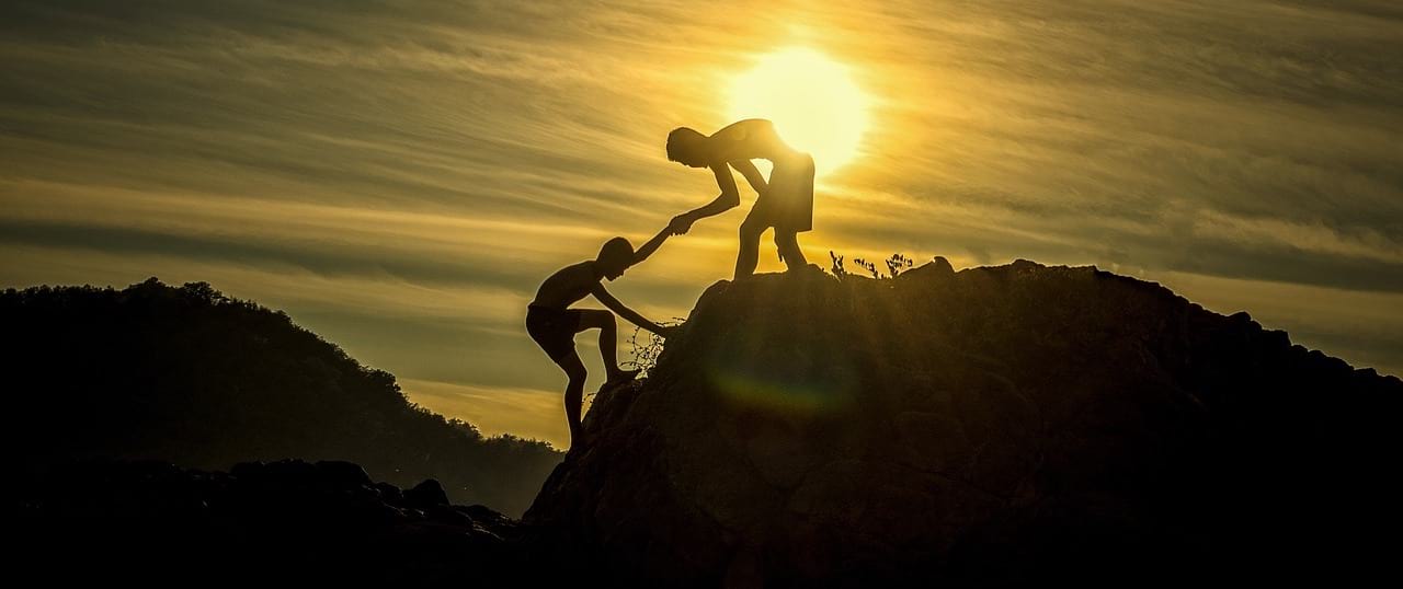Two people climbing to the top of a mountain. The person in front is helping the person behind.